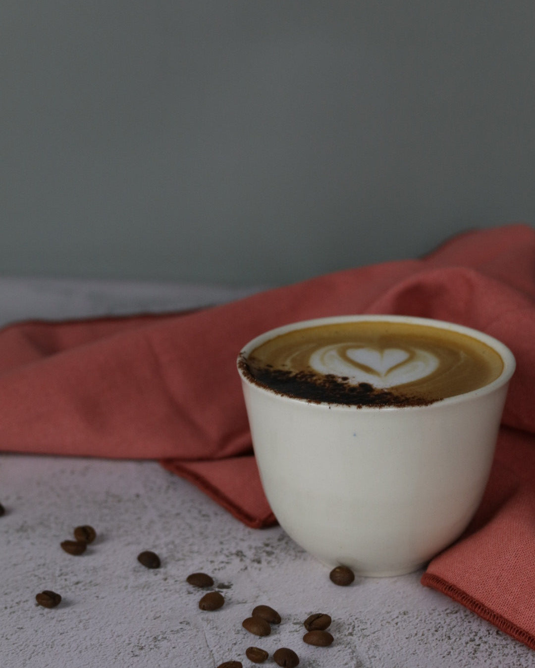 White ceramic cup of coffee on a pink napkin with coffee beans scattered around