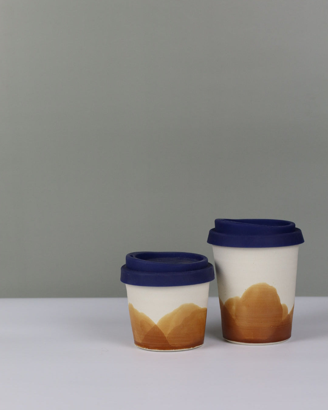 Small and large ceramic travel tumbler with ochre glaze and blue silicon lids on a grey background.