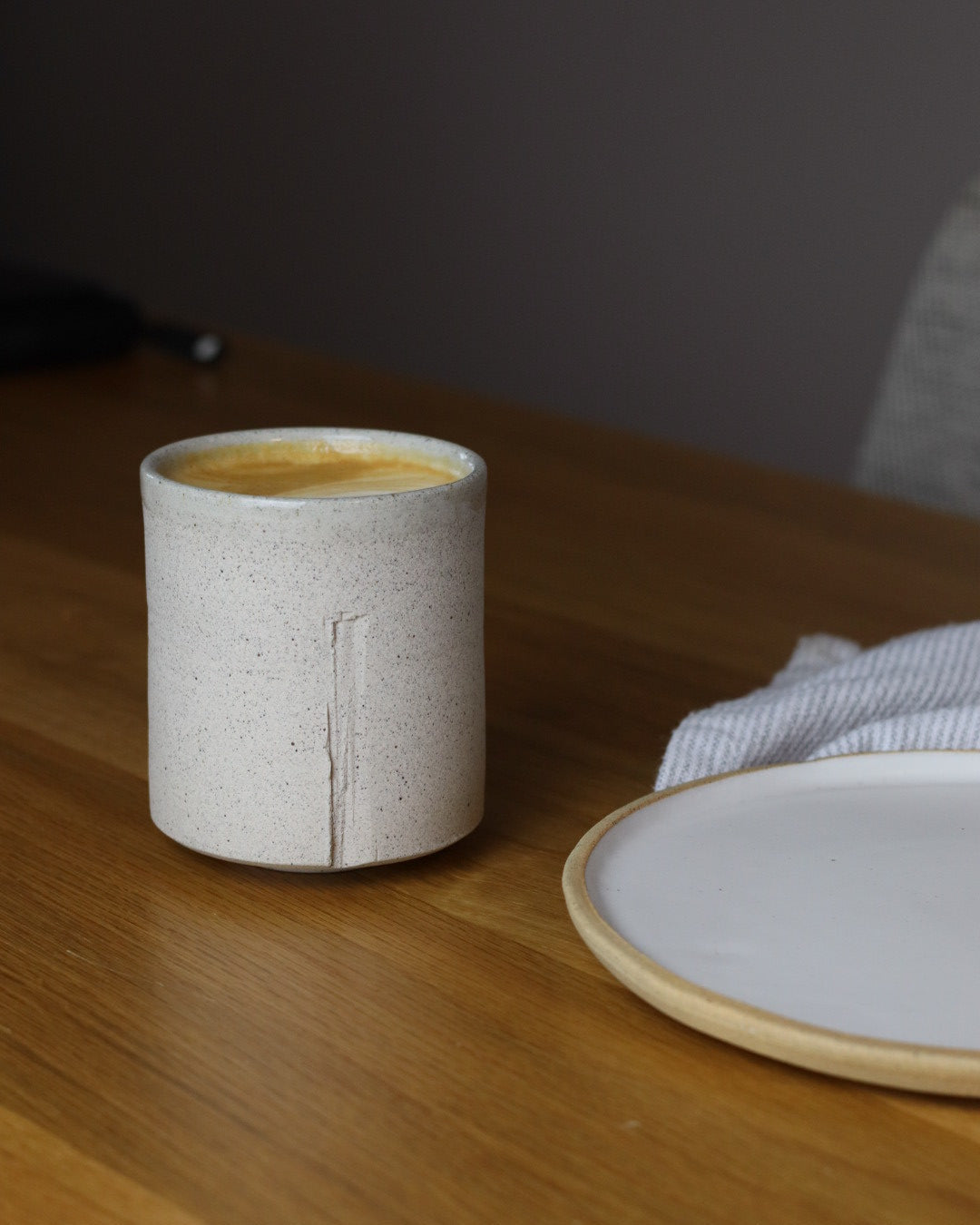 Ceramic tumbler of coffee next to a white ceramic plate on a wooden table