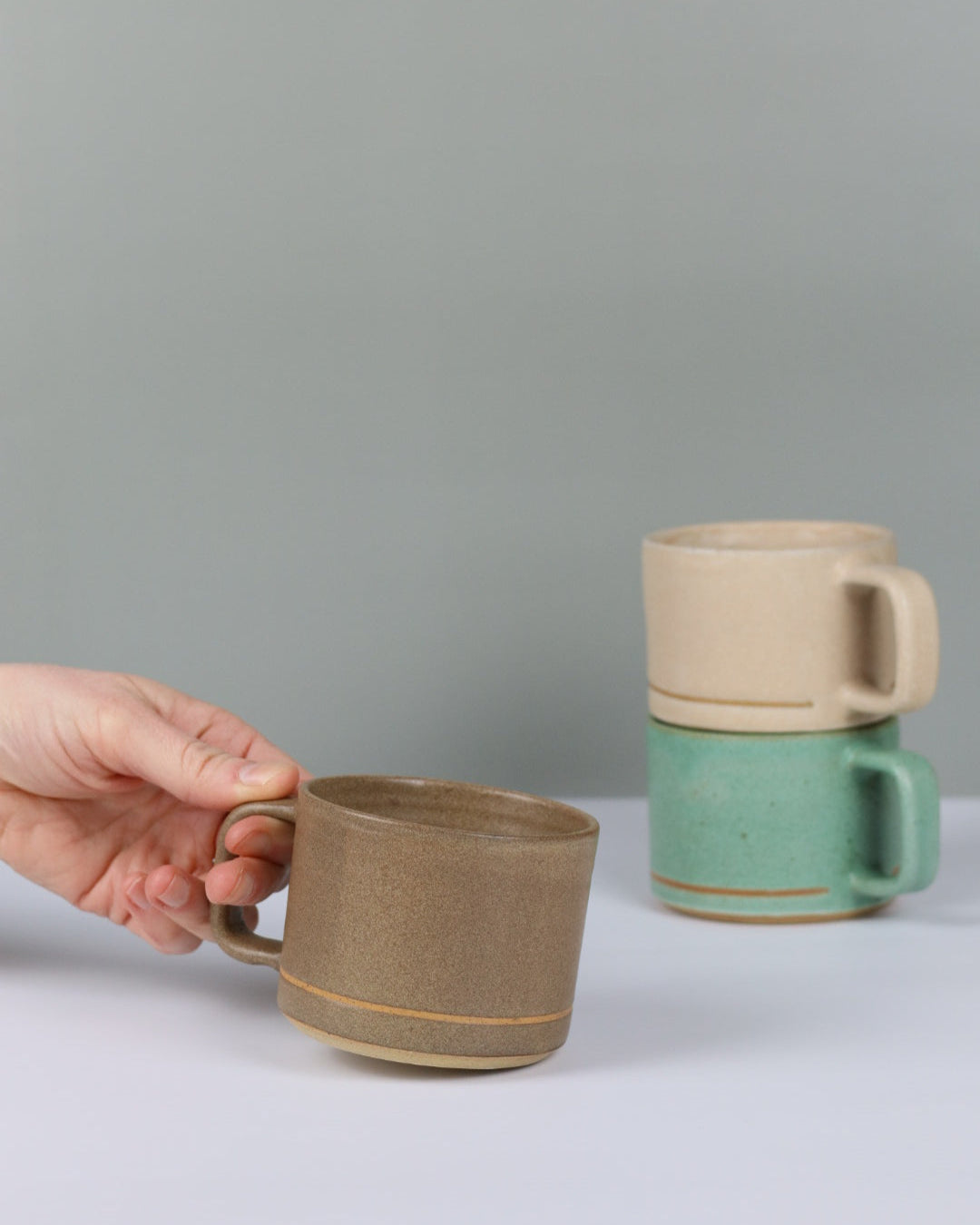 Three ceramic cups with handles, one brown, one green, and one oatmeal held against a plain background.