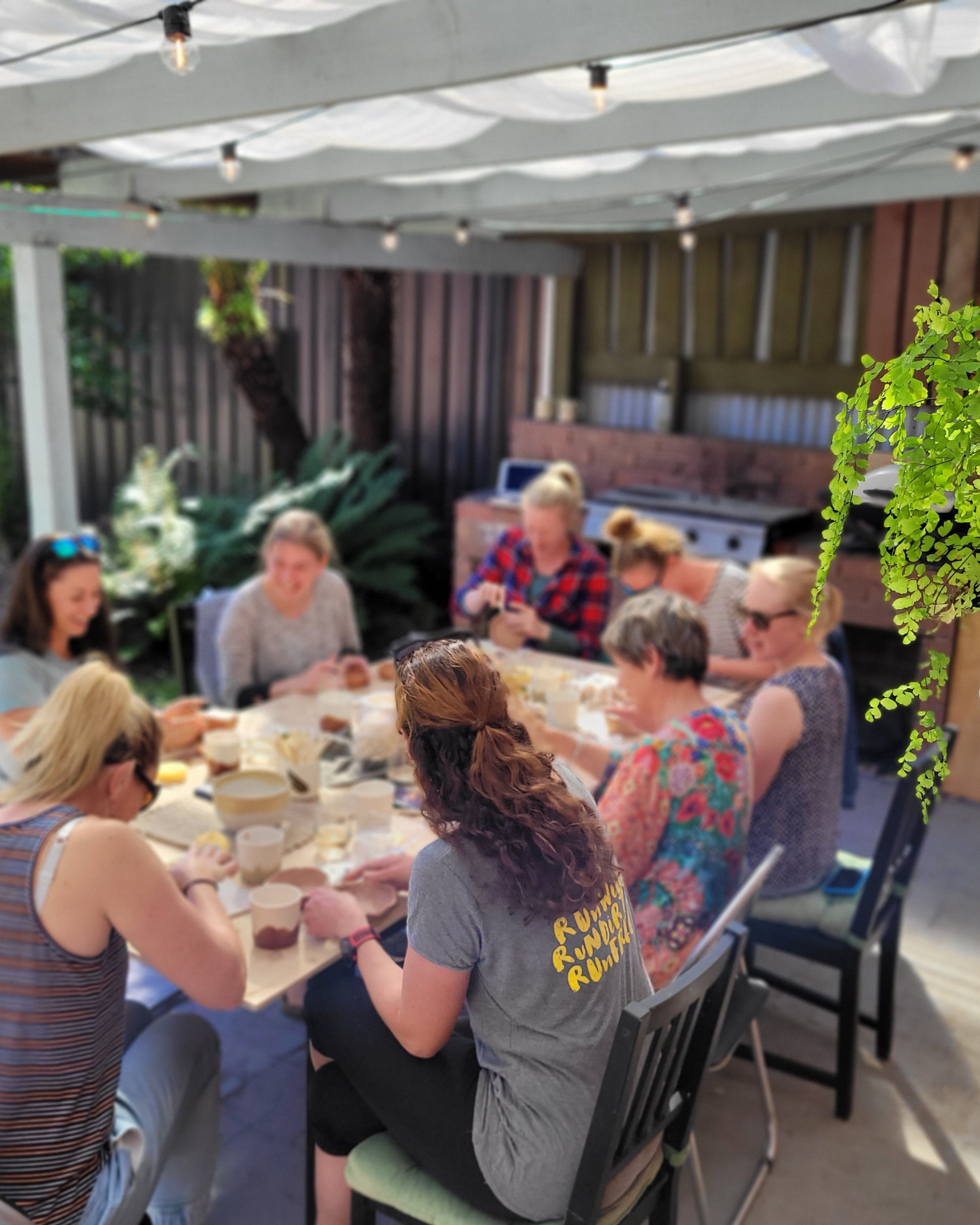 Group of people sitting around a table outdoors under a pergola working with clay.