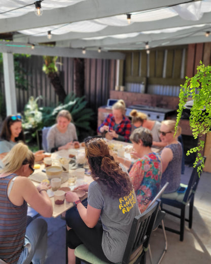 Group of people sitting around a table outdoors under a pergola working with clay.