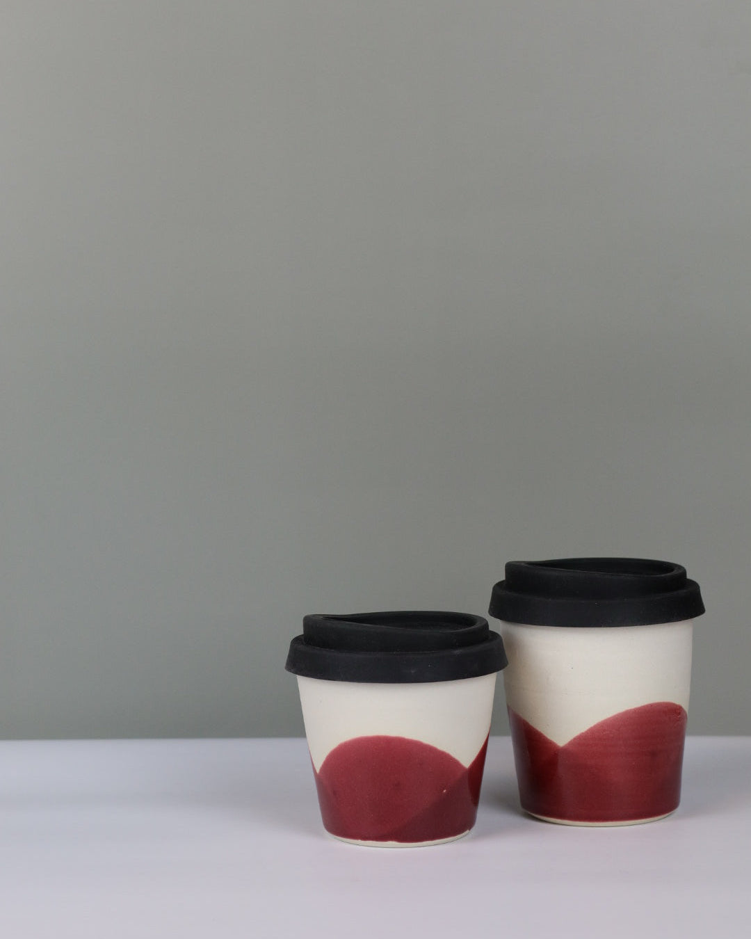 Small and large ceramic travel tumblers with cranberry red glaze and black silicon lids on a grey background.