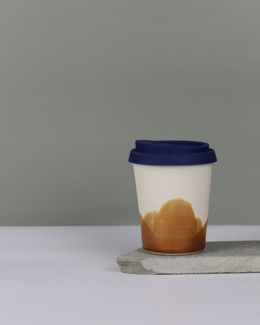 Ceramic travel tumbler with ochre glaze and blue silicon lid on a grey background.