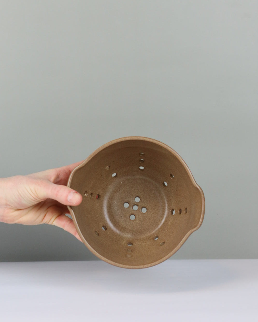 Hand holding a brown ceramic strainer against a plain background
