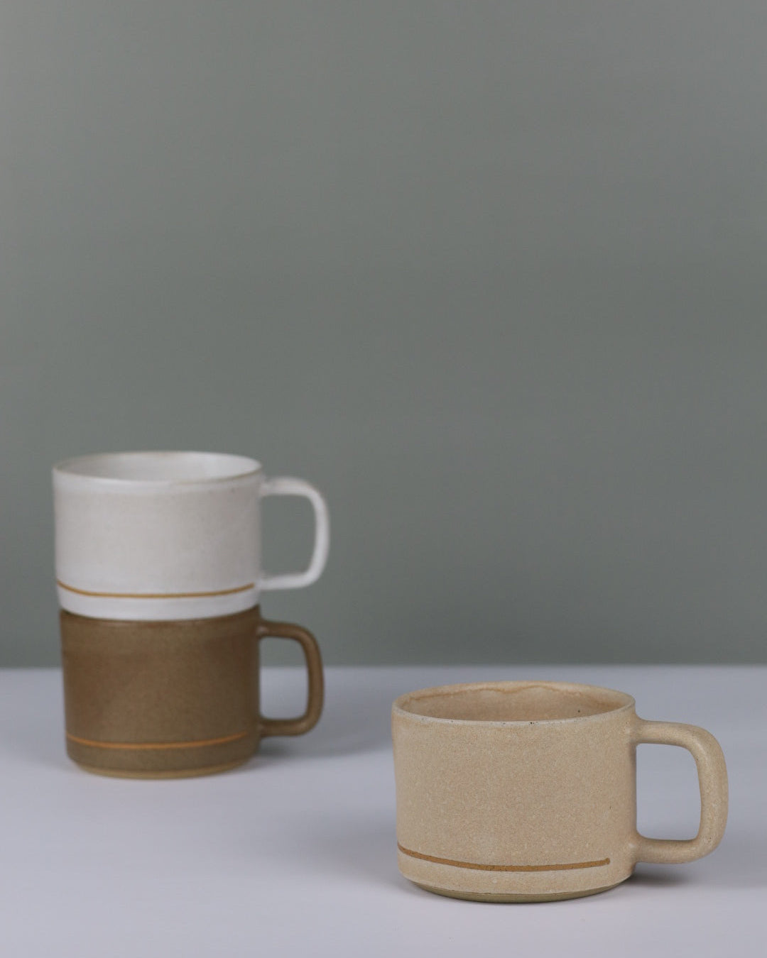 Three ceramic mugs in beige, brown, and white on a grey background.