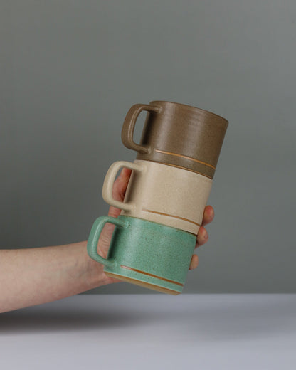 Hand holding three ceramic mugs, one green, one oatmeal and one brown, against a grey background