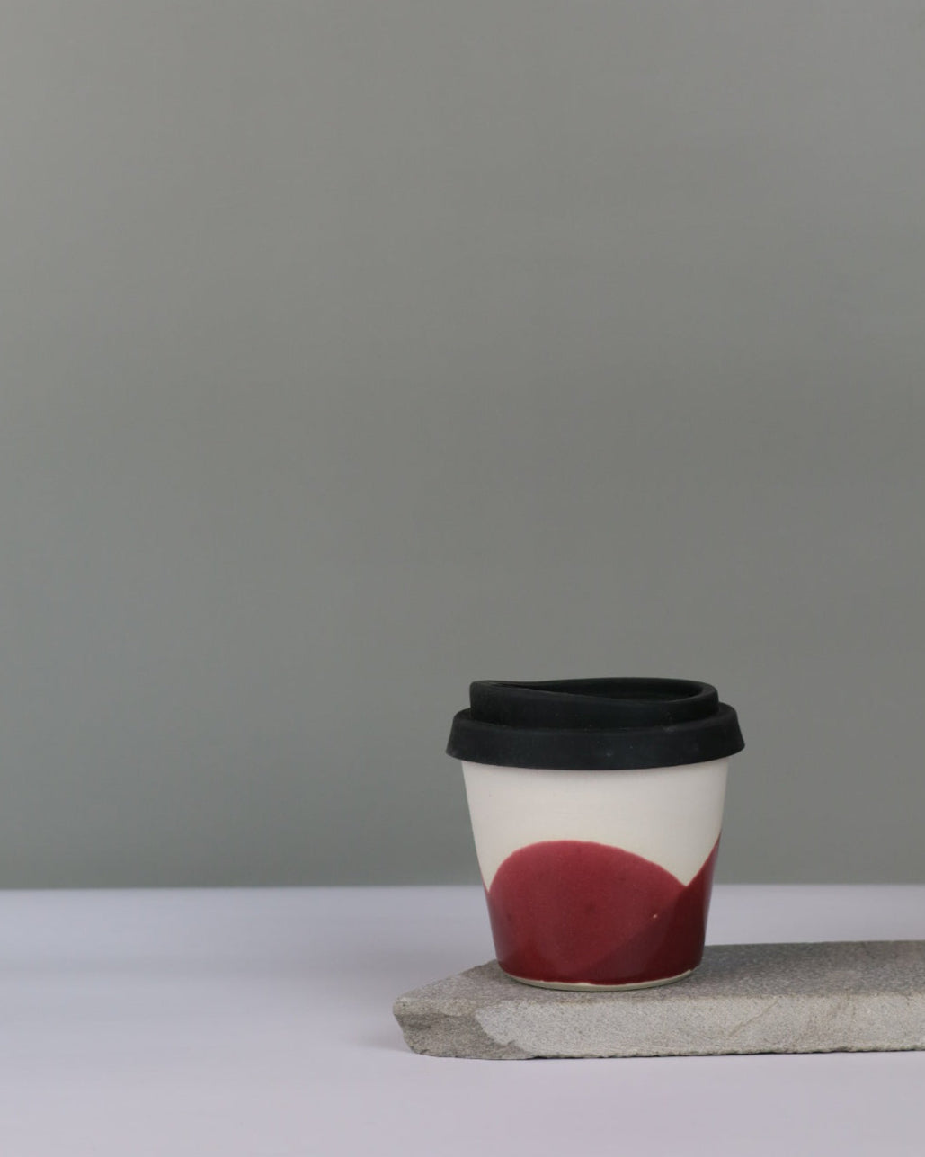 Ceramic coffee cup with a red glaze and a black lid on a grey background