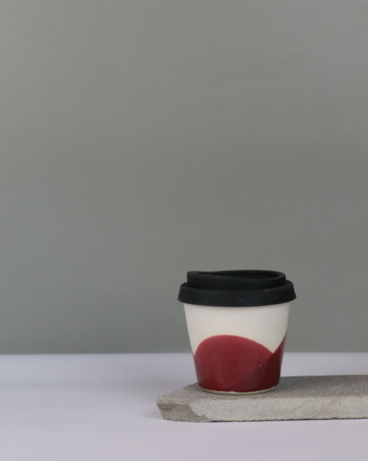 Ceramic coffee cup with a red glaze and a black lid on a grey background
