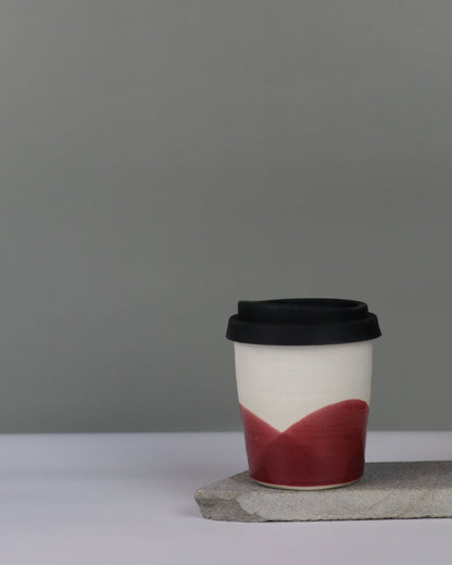 Ceramic travel tumbler with cranberry red glae and black silicon lid on a grey background.