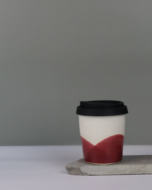 Ceramic travel tumbler with cranberry red glae and black silicon lid on a grey background.